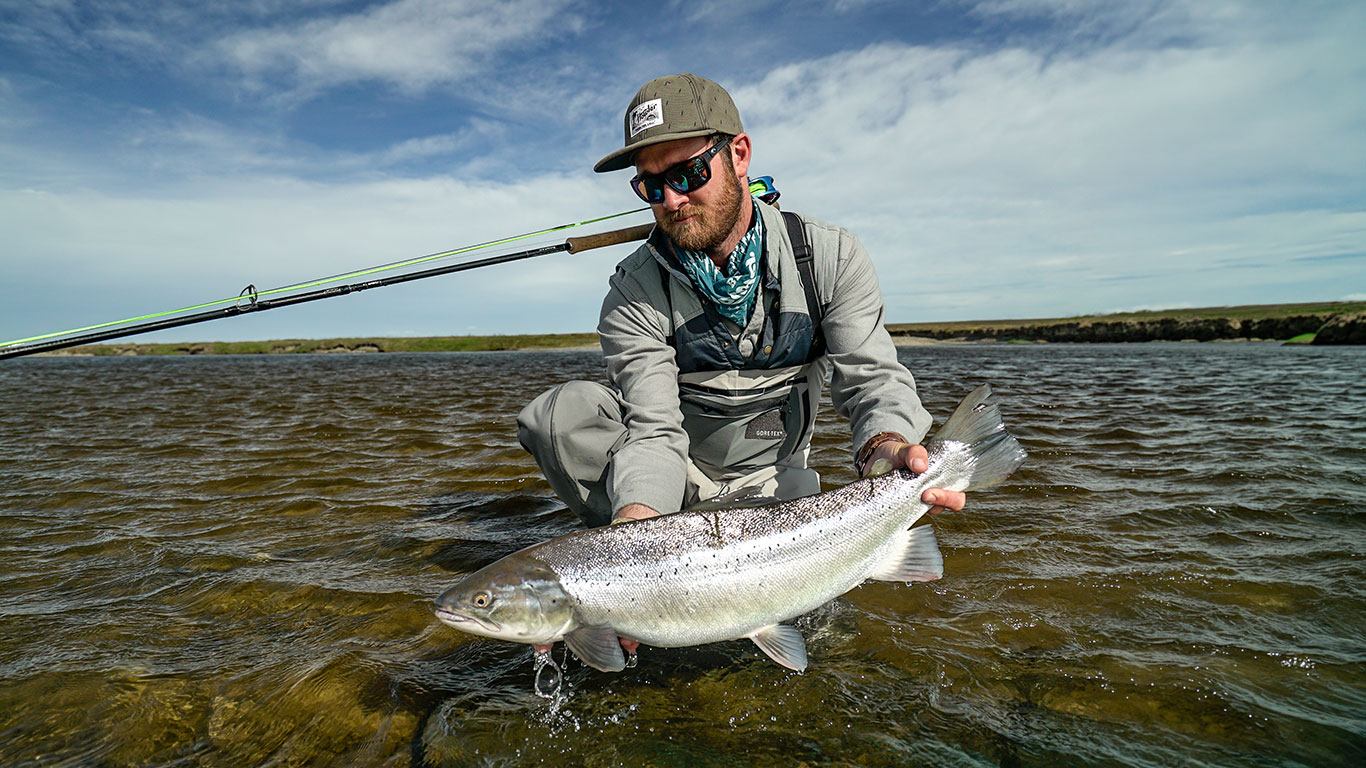 Angler kneeling in shallow water, holding a large silver fish with a fishing rod over his shoulder.