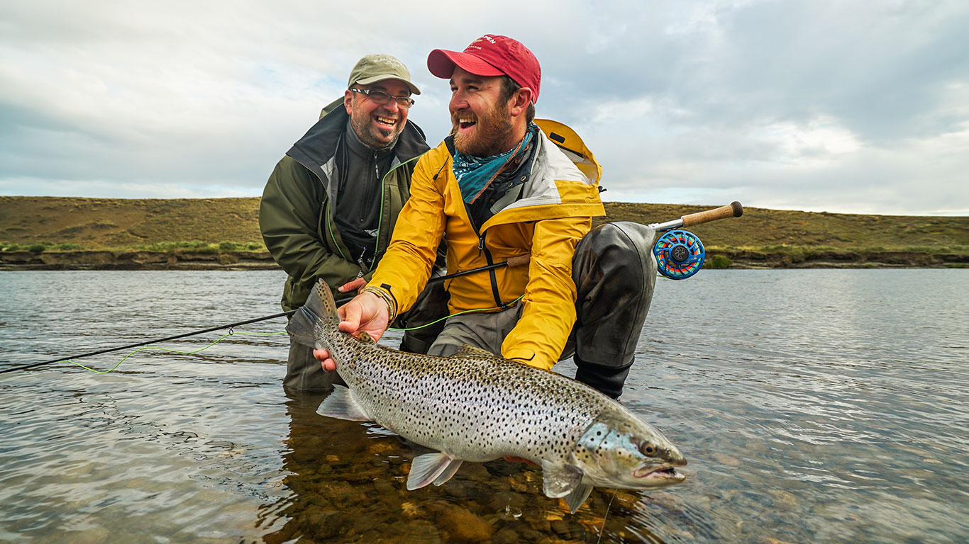 Two fishermen kneel in a shallow river, proudly holding a large speckled trout they just caught.