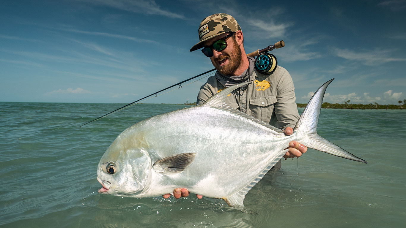 Man in a camouflage hat and sunglasses holds a large silver fish in shallow turquoise water.