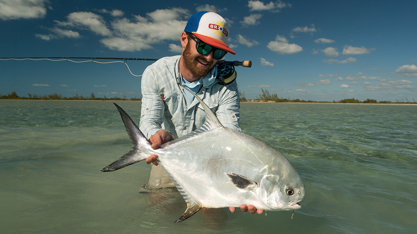 Smiling fisherman in a cap and sunglasses holds a large silver fish in waist-deep, clear blue-green water under a sunny sky.