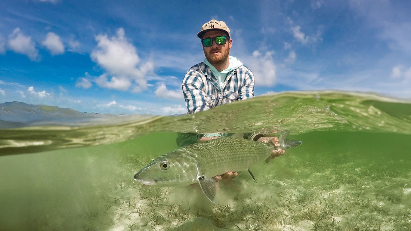 Person wearing a cap and sunglasses holds a large fish underwater, with clear blue sky above and shallow seagrass below the surface (split view).