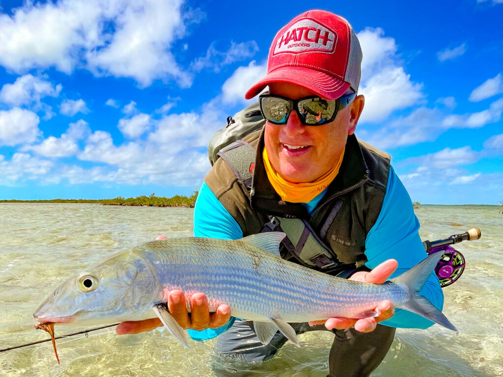 Fisherperson in a red cap and sunglasses holding a large silver fish at the shoreline with a blue sky and shallow water behind them.