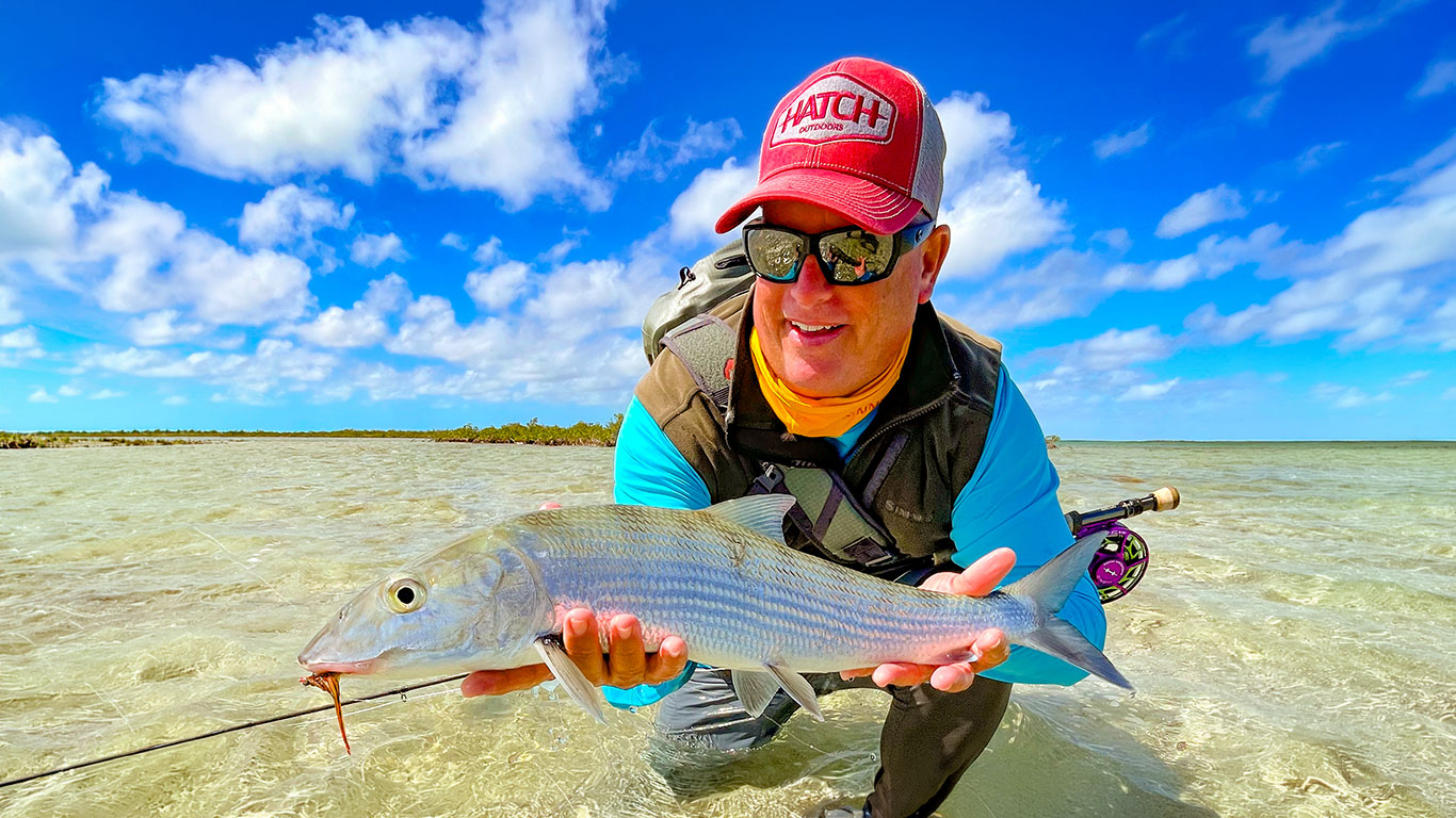 Angler in a red cap and sunglasses holds up a large silvery fish in shallow, clear tropical water with a bright blue sky behind him.