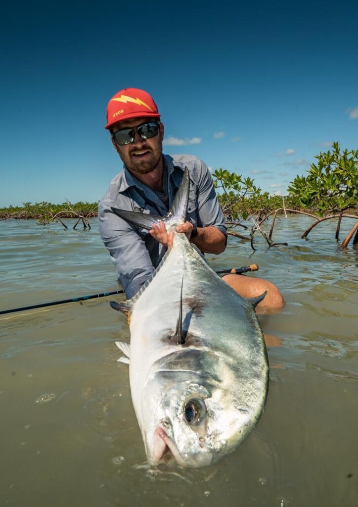 Man in sunglasses and a red cap holds a large silver fish in shallow water, with mangrove shrubs in the background under a clear blue sky.