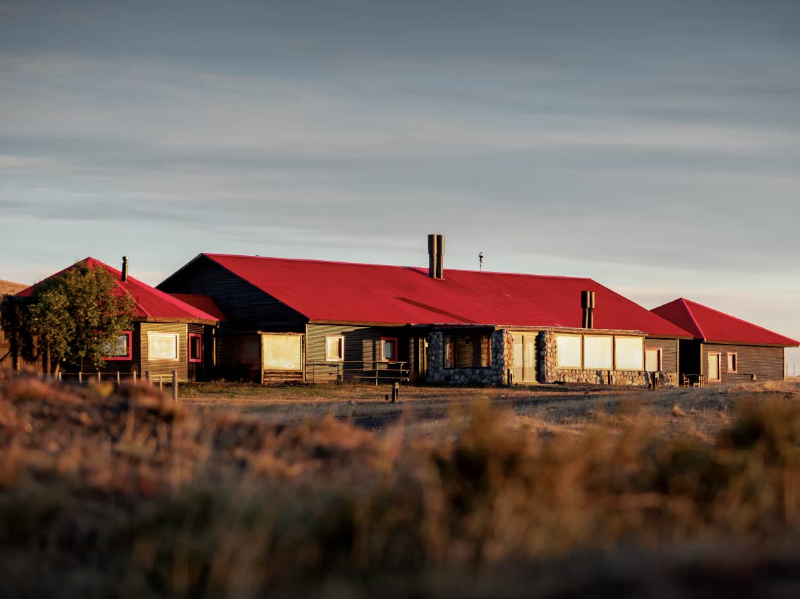 Rustic, single-story lodge with a bright red roof set in a dry grassland at sunset.