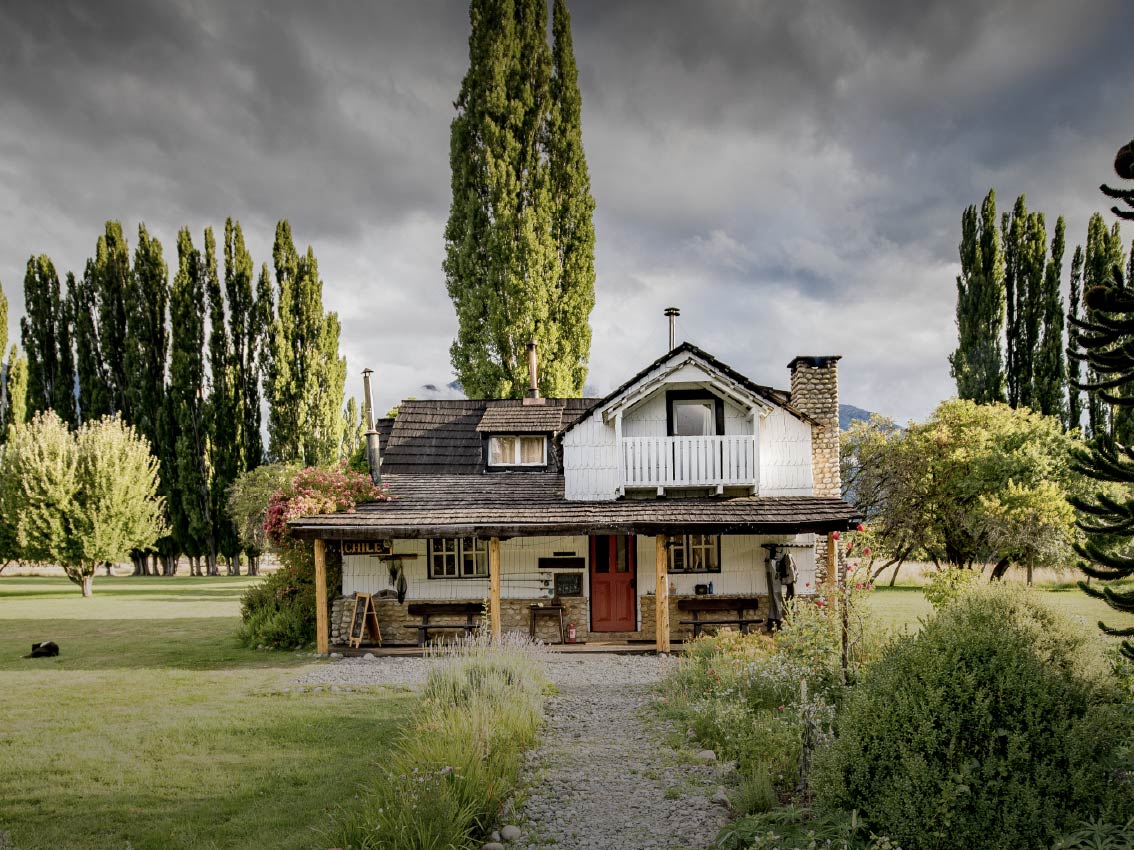 Rustic two-story cottage with a red front door, white balcony, and a garden under a cloudy sky.